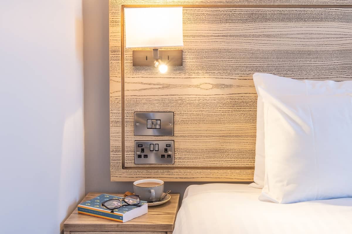 A neatly made bed with white pillows, a bedside table with a book, glasses, and a cup of coffee, and a wall-mounted light fixture.