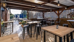 An outdoor patio area with wooden tables and chairs, a brick wall, and string lights. - Two Rivers, Chepstow by Marston’s Inns (Chepstow)