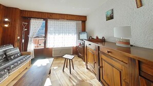 L’intérieur d’un chalet accueillant, avec un banc en bois, une petite table, un coin cuisine doté de placards, un téléviseur et une fenêtre avec des rideaux.