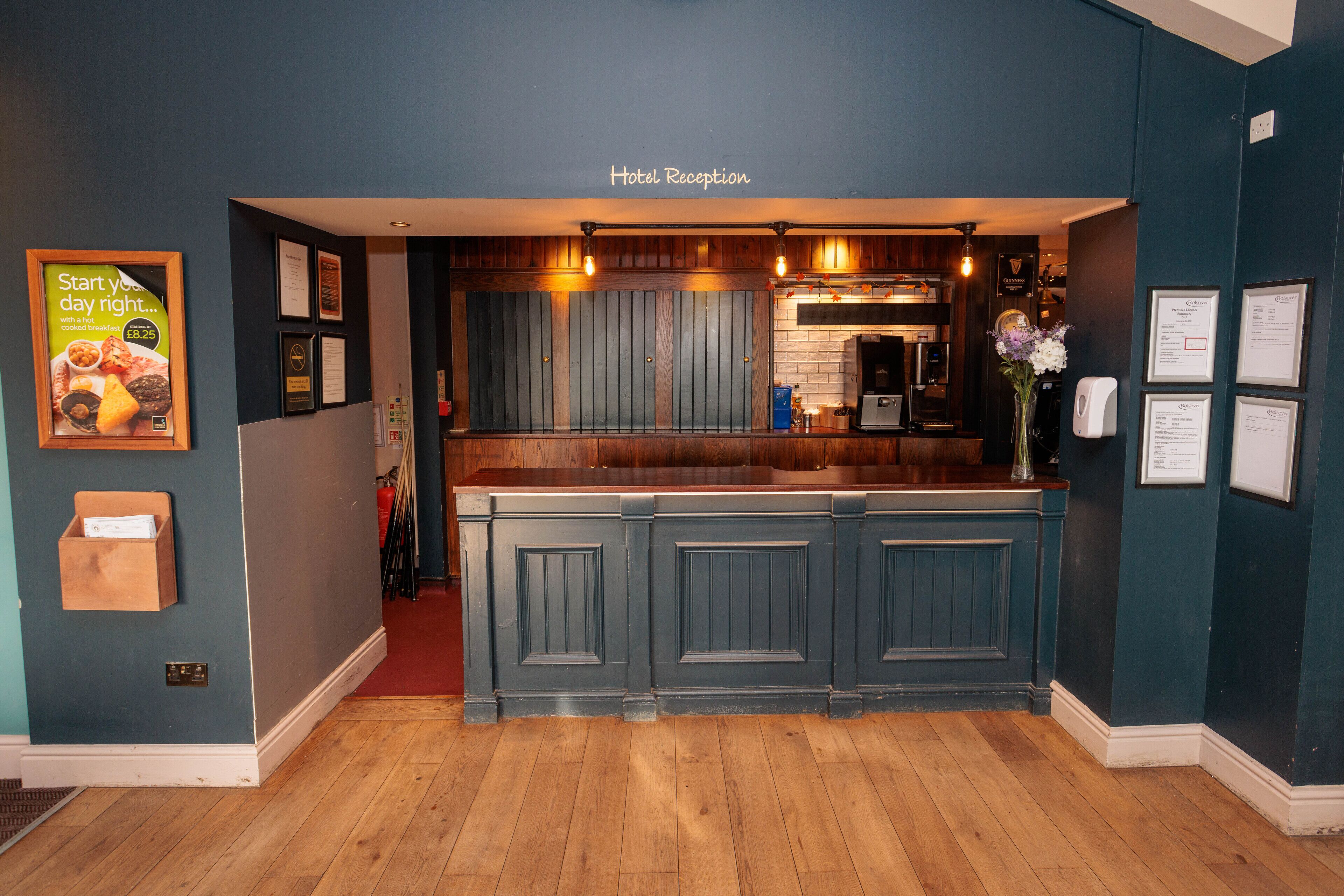 A hotel reception area with a wooden counter, framed certificates, and a sign above the counter reading "Hotel Reception".