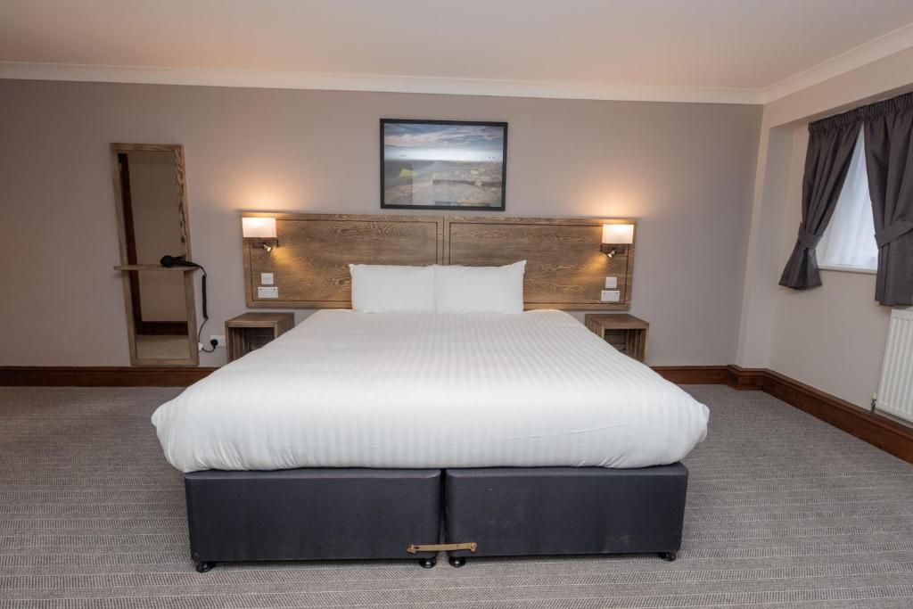 A hotel room with a large bed, two bedside tables, a headboard with wall-mounted lamps, a framed picture above the bed, and a window with curtains.