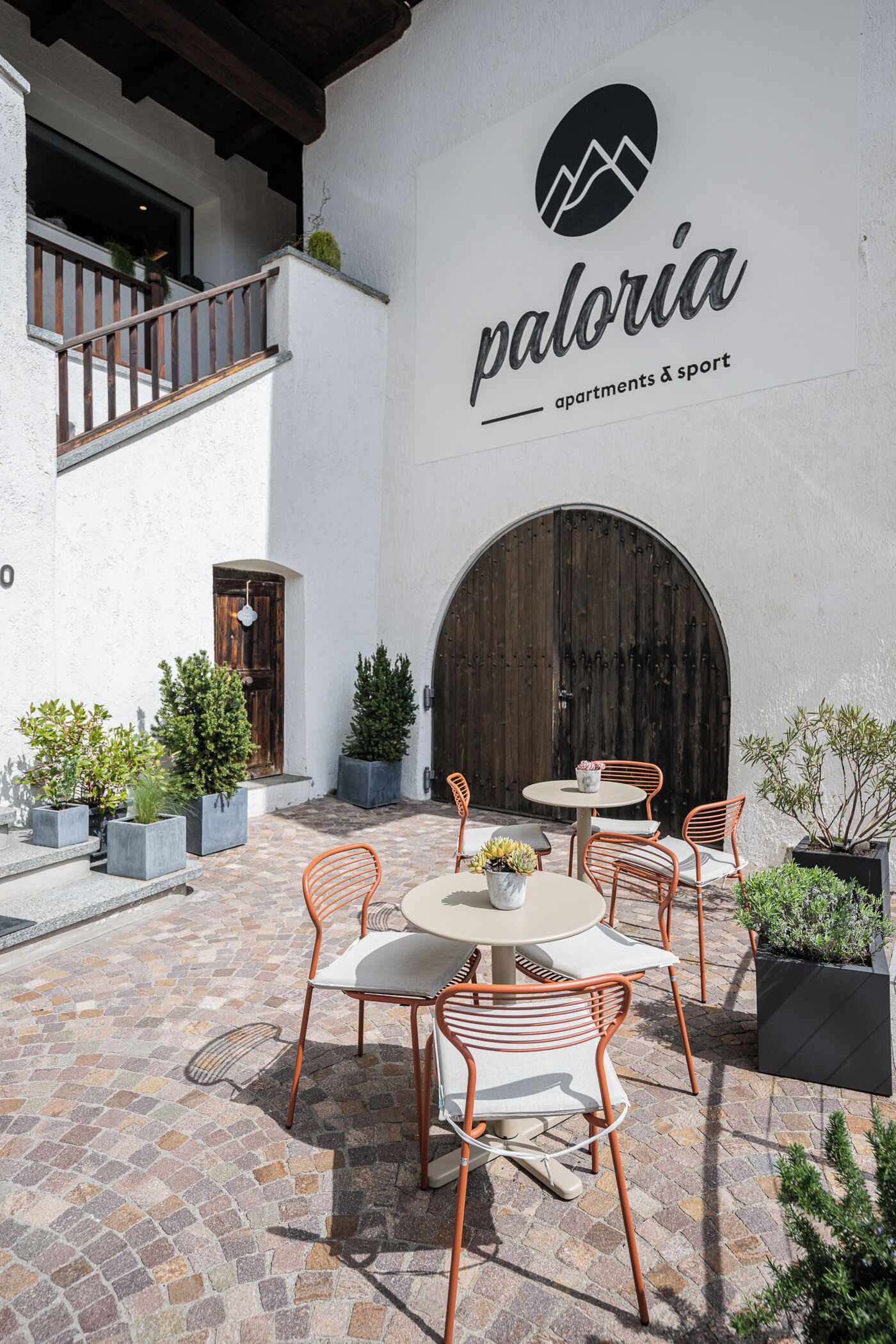 Un espacio de patio al aire libre con una mesa redonda y sillas naranjas, plantas en macetas y una puerta de madera grande.