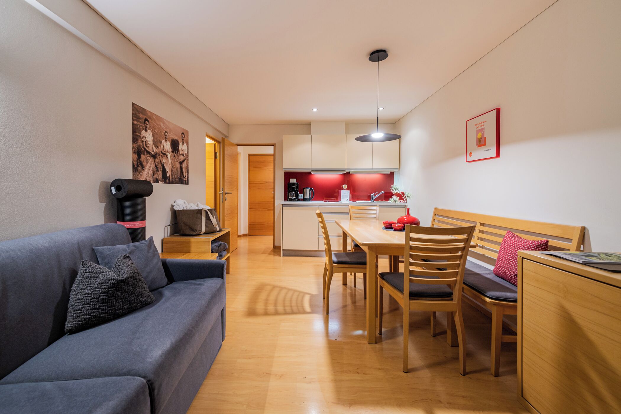 A modern living room with a grey sofa, wooden dining table, and chairs. A kitchen with red accents is visible in the background.