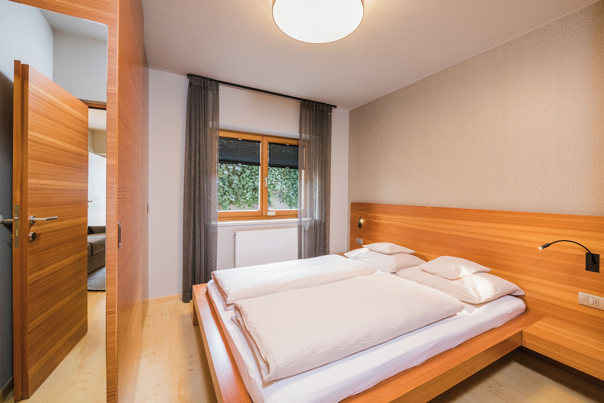 A bedroom with a wooden bed, a window with curtains, and a door leading to another room.
