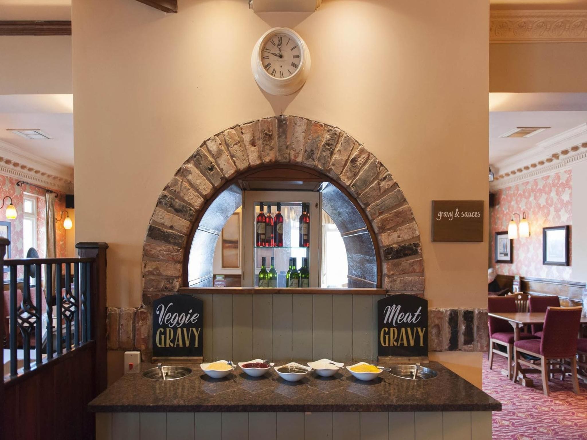 Mostrador de restaurante con un arco de ladrillo, un reloj encima y letreros que dicen 'Salsa de Verduras' y 'Salsa de Carne' a cada lado.