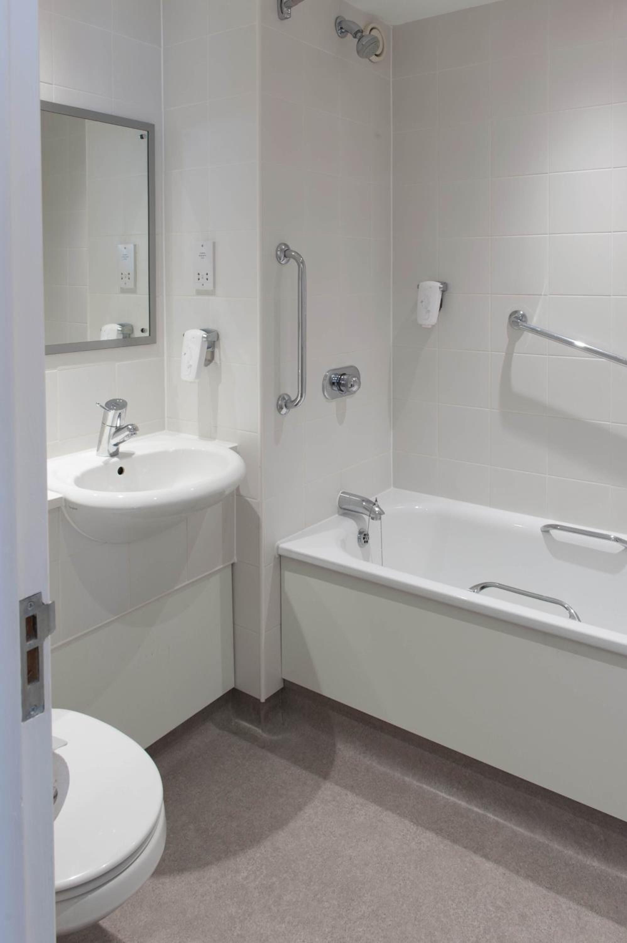 Un baño con lavamanos, inodoro y bañera blancos. Cuenta con barras de apoyo y un perchero.