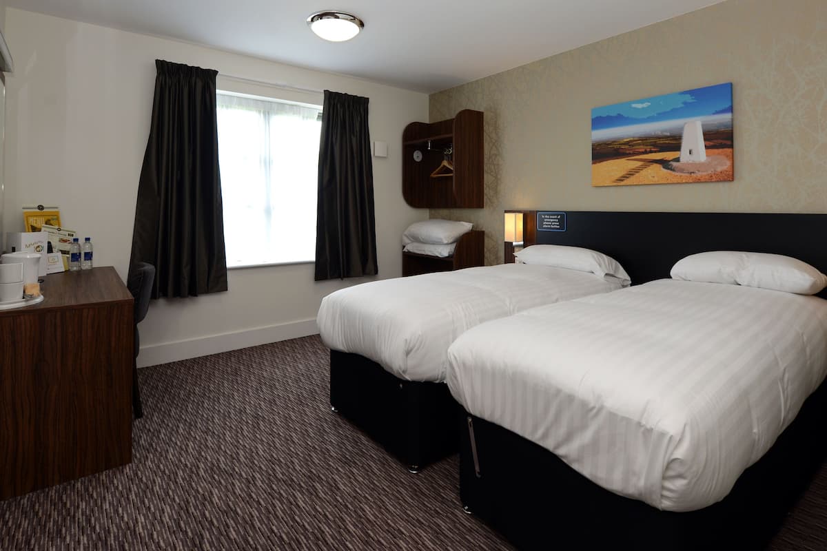 A hotel room with two beds, a desk, a coat rack, and a painting on the wall.