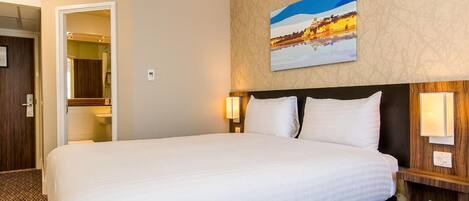A hotel room with a large bed, two bedside lamps, a painting on the wall, and a door leading to a bathroom.