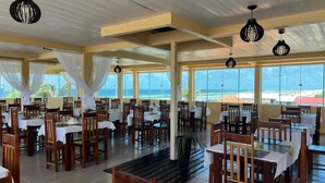 A restaurant with multiple tables and chairs, large windows with ocean views, and hanging pendant lights. - Hotel Concha do Mar (Salinópolis)