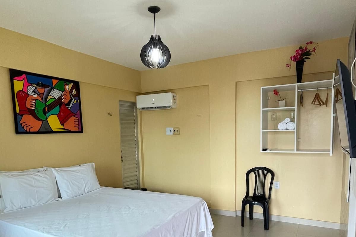 Quarto com cama, um quadro, ar condicionado, uma cadeira e uma estante com objetos.