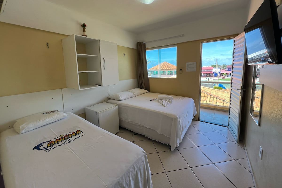 Quarto de hotel com duas camas, um pequeno armário branco, uma televisão e vista para o exterior.