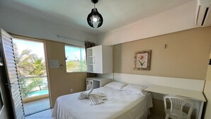 A hotel room with a bed, a desk, a chair, a wardrobe, a small refrigerator, a window with a view of palm trees, and a ceiling light. - Hotel Concha do Mar (Salinópolis)