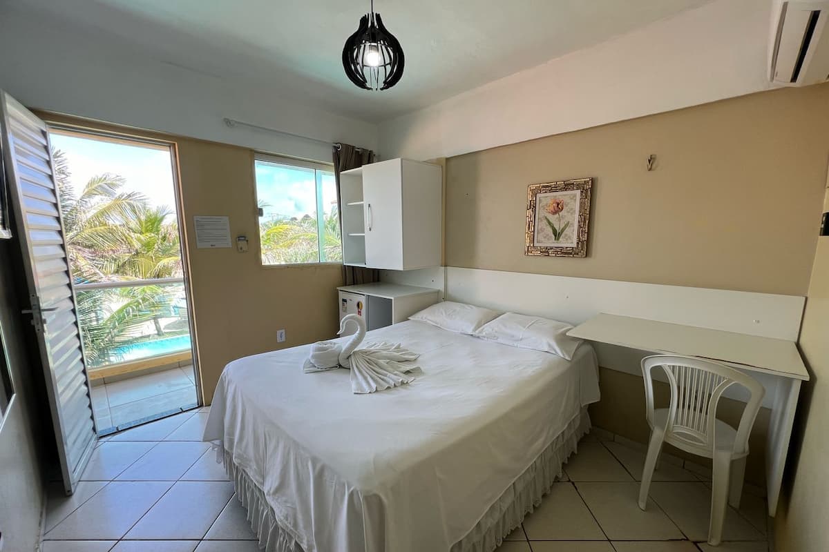 Quarto de hotel com cama, escrivaninha, cadeira, guarda-roupa, pequeno frigorífico, janela com vista para palmeiras e luz de teto.