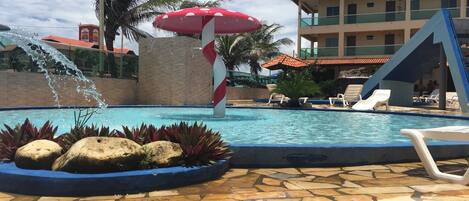 Una piscina con fuente, sillas de descanso y un edificio con techo de tejas al fondo.