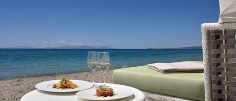 Una mesa de comedor con dos platos de comida y dos copas de vino blanco, ubicada en una playa con vista al mar y montañas lejanas.