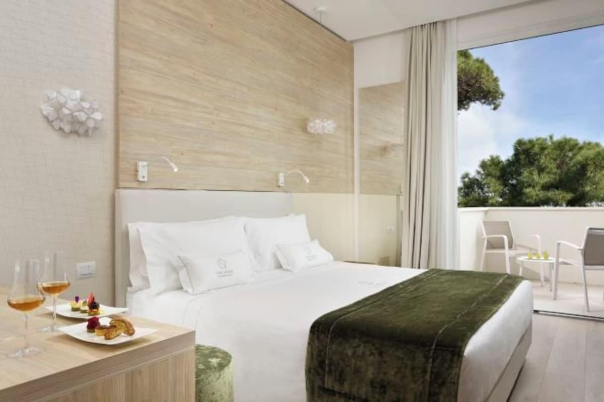 A modern hotel room with a bed, a bedside table with glasses and snacks, a view of trees through the window, and a balcony with chairs.