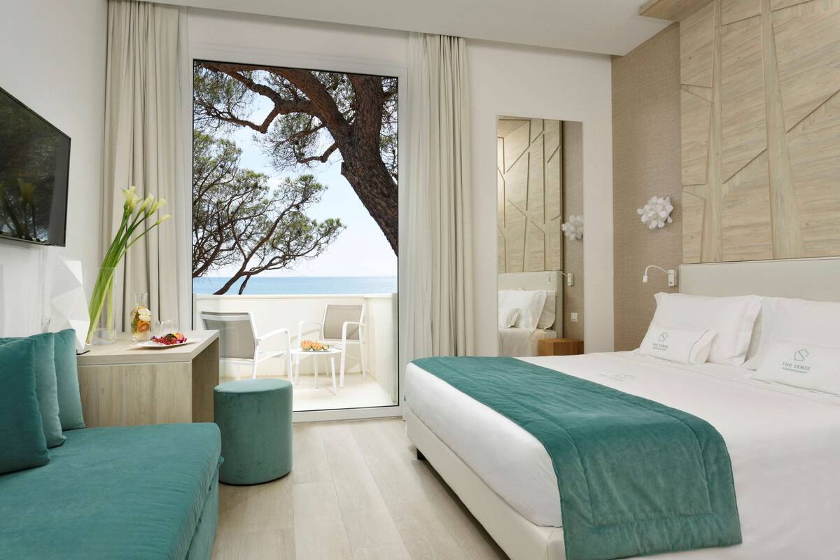 A hotel room with a large bed, a desk with a chair, a small round ottoman, and a view of the sea through a large window.