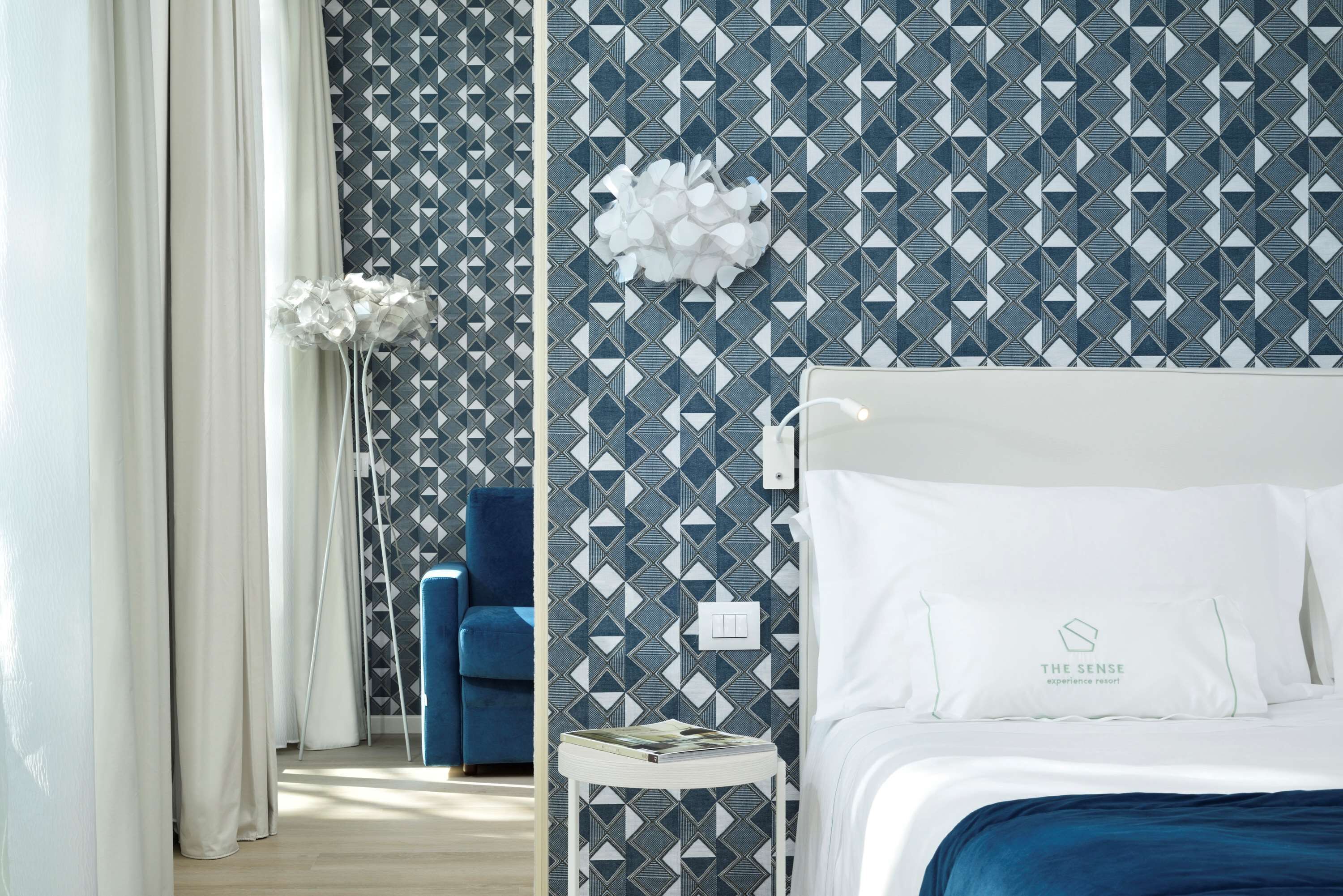 A hotel room with a geometric patterned wallpaper, a blue sofa, a white bedside table with a magazine, and a bed with white bedding.