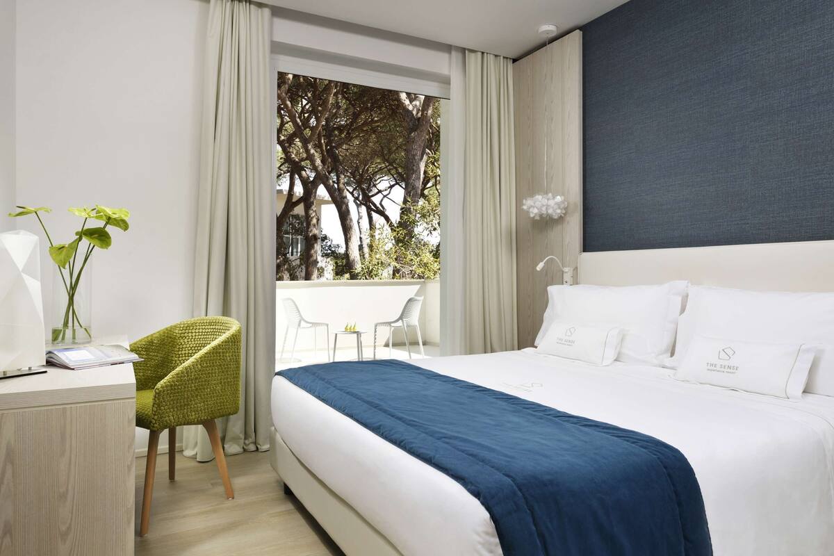 A hotel room with a large bed, a desk with a chair, a window with curtains, and a view of trees outside.