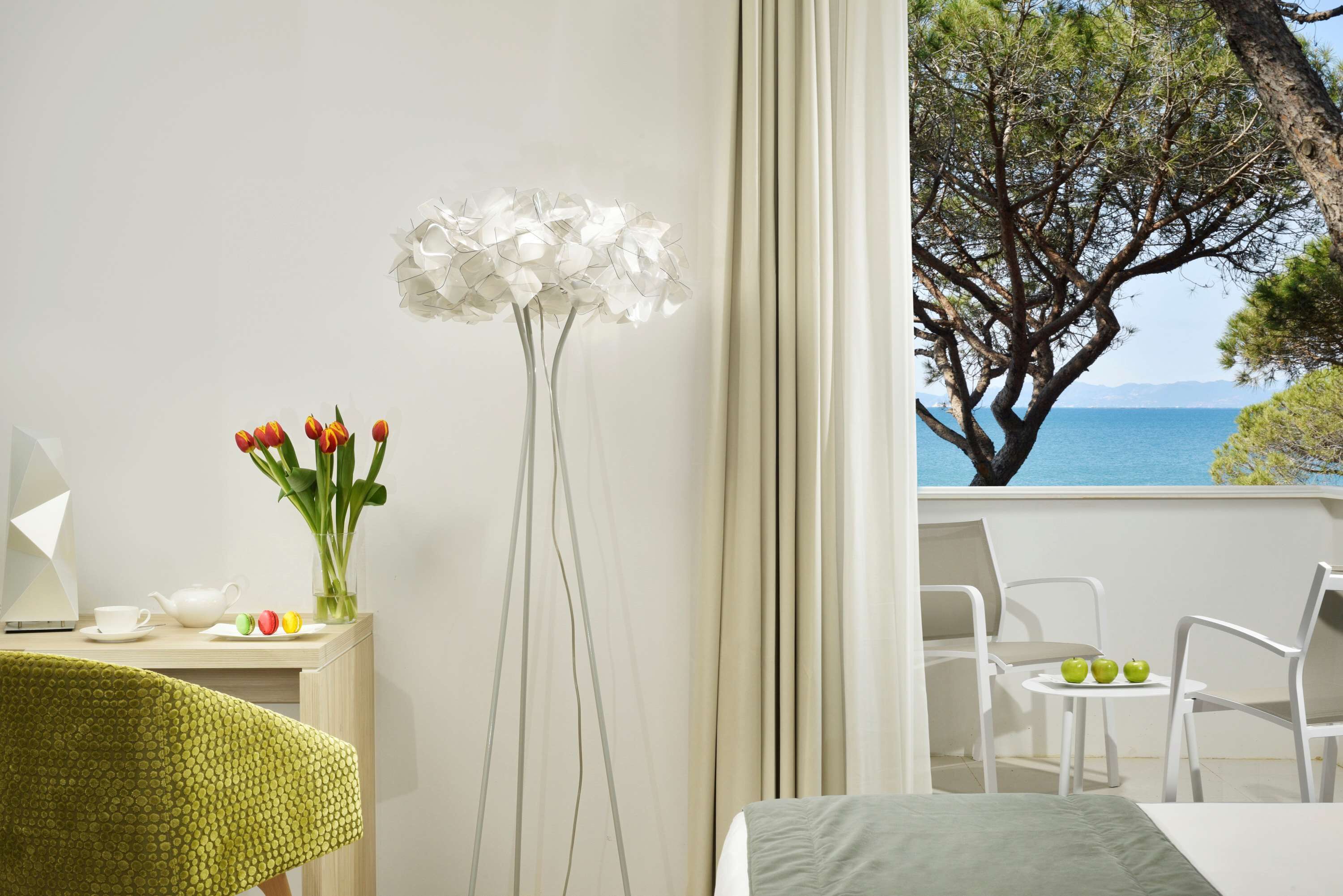 A hotel room with a balcony, a vase with flowers, a table with apples, and a view of the sea.