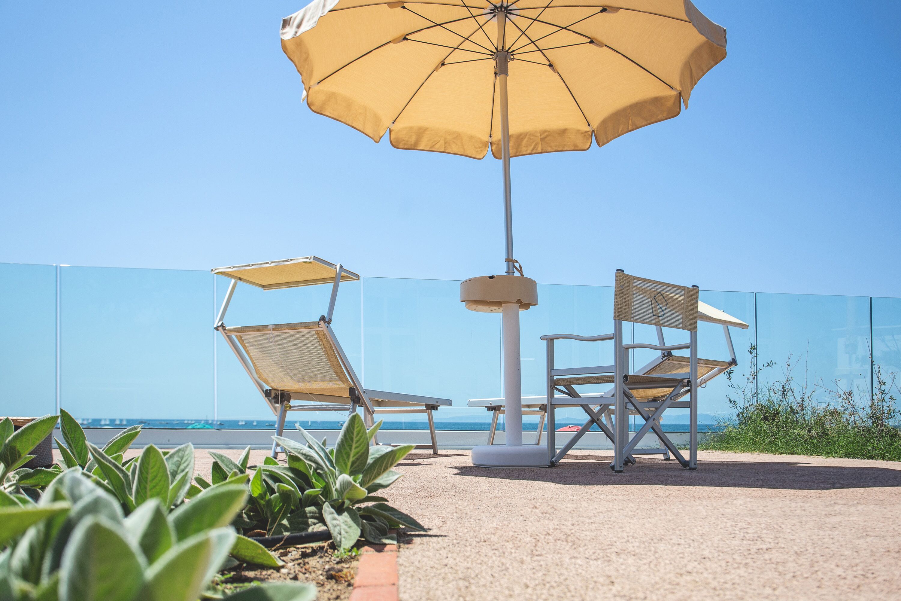 Un patio frente a la playa con dos tumbonas y una mesa bajo un paraguas grande, con vista al mar.