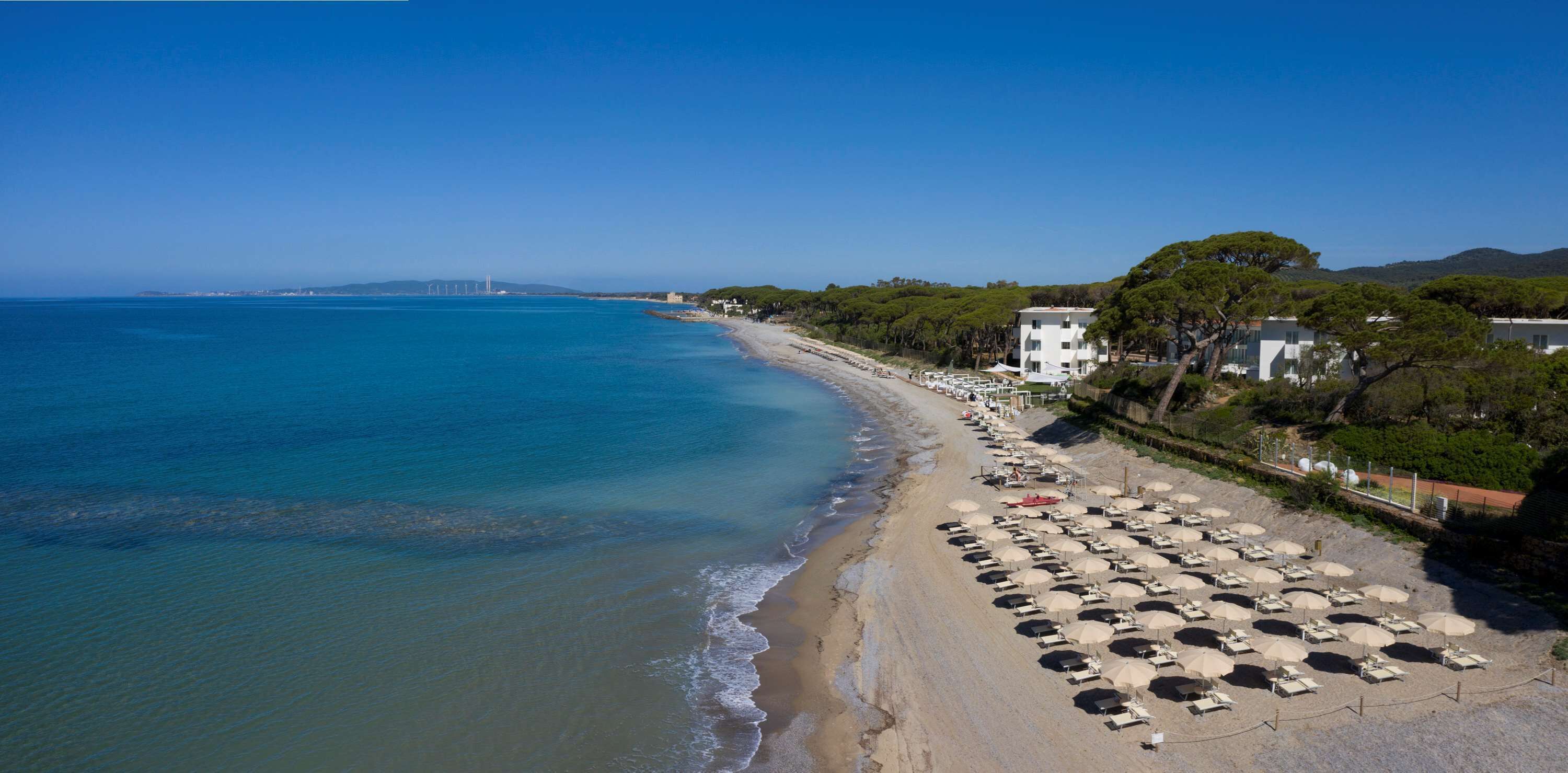 A beach with rows of sunbeds and umbrellas, a clear blue sea, and a bridge in the distance.