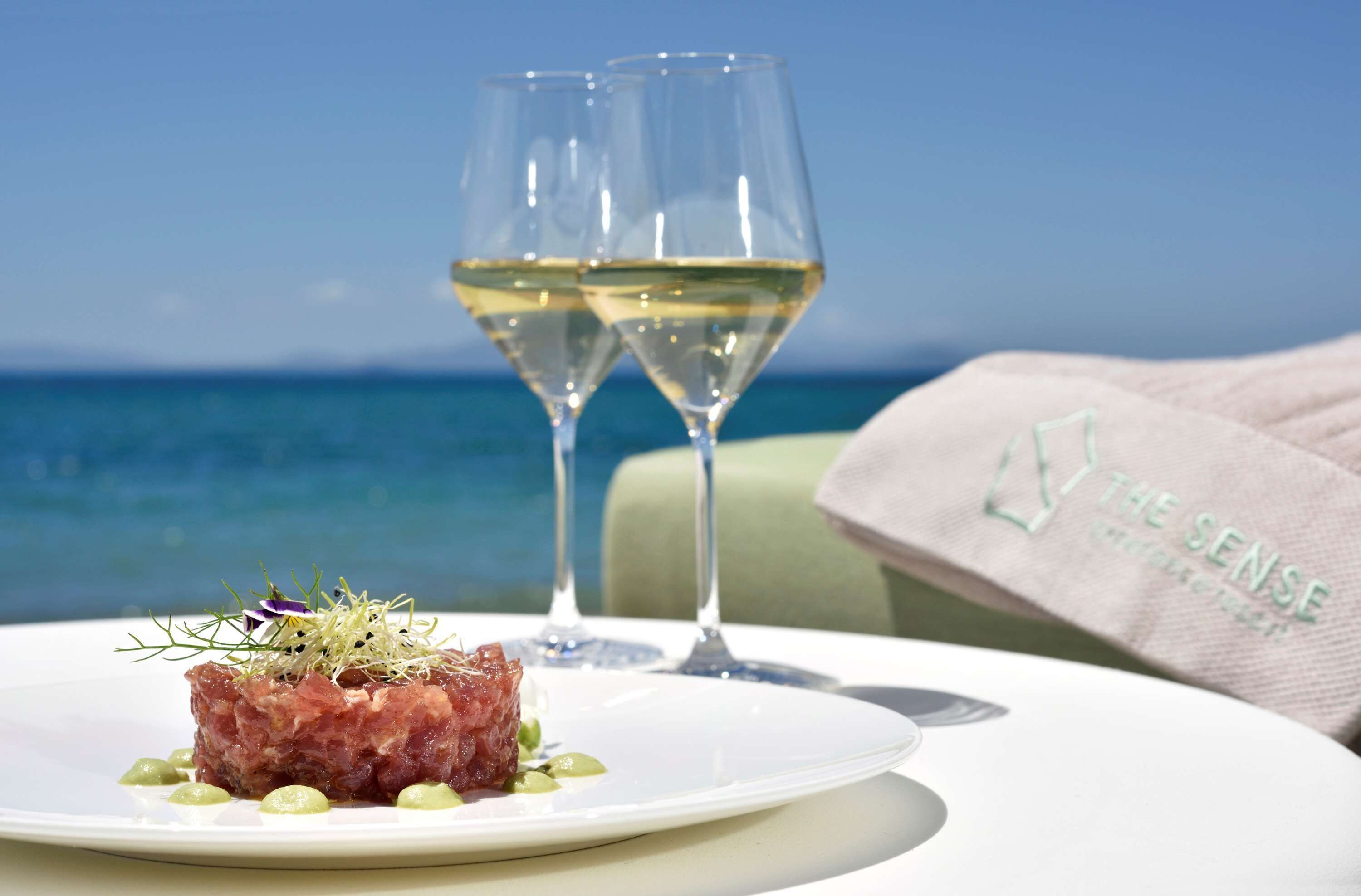 Une assiette de nourriture, accompagnée d’un verre de vin blanc et d’une serviette en papier avec du texte, le tout présenté sur un fond de ciel bleu et d’océan.