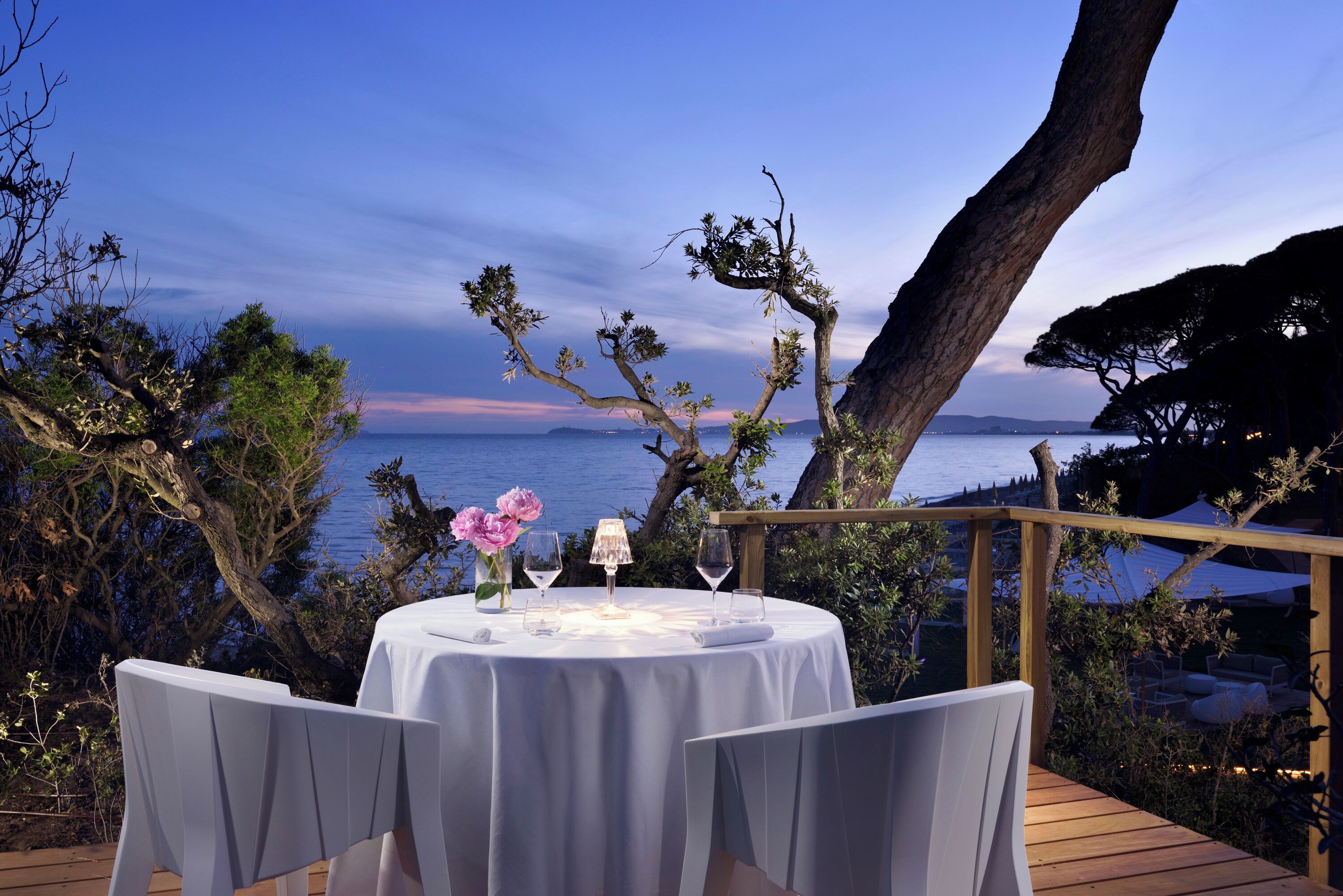 Mesa redonda con mantel blanco, dos copas de vino y flores rosadas sobre una terraza de madera con vista a un cuerpo de agua.