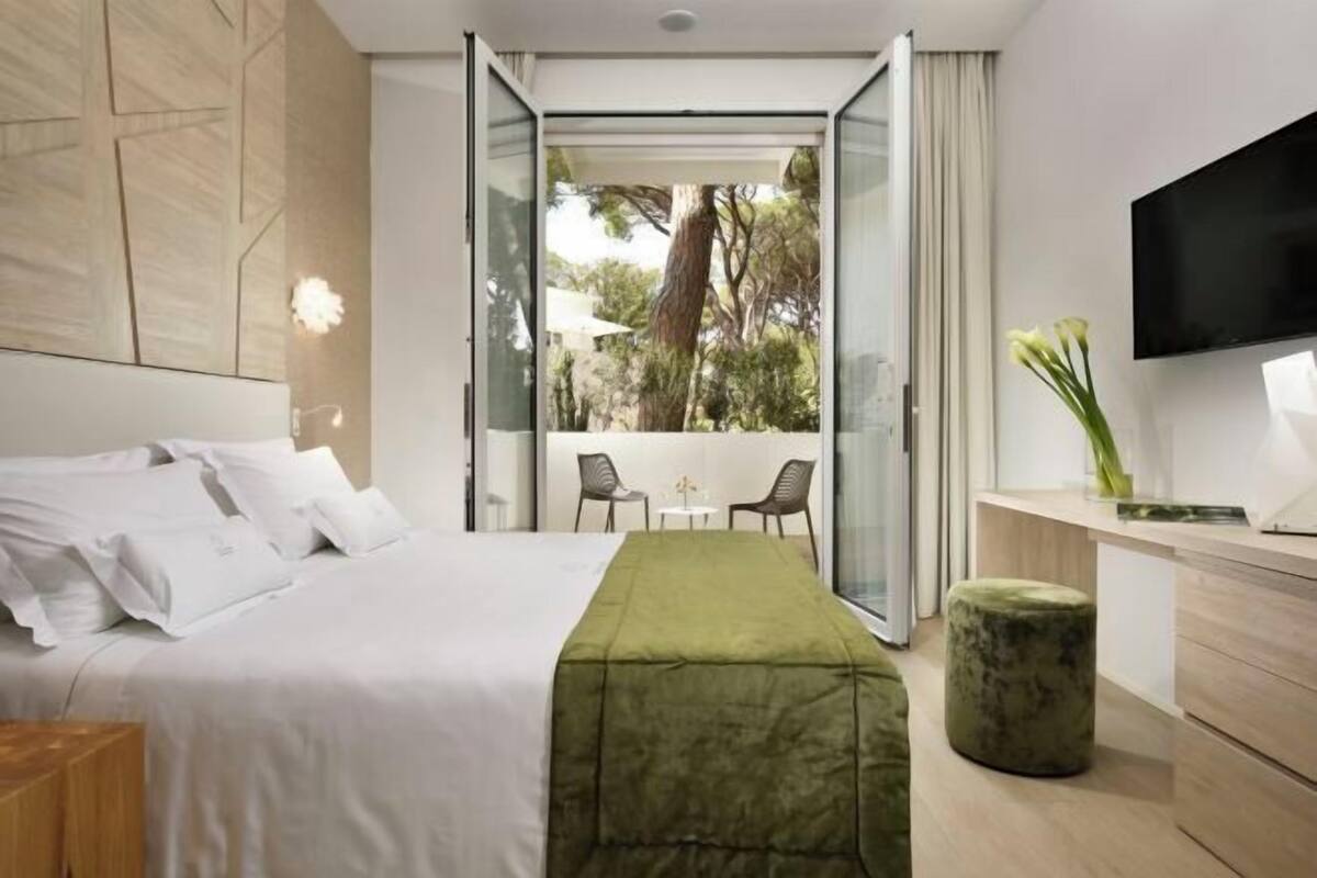 A modern hotel room with a bed, a desk, a TV, and a balcony with chairs.