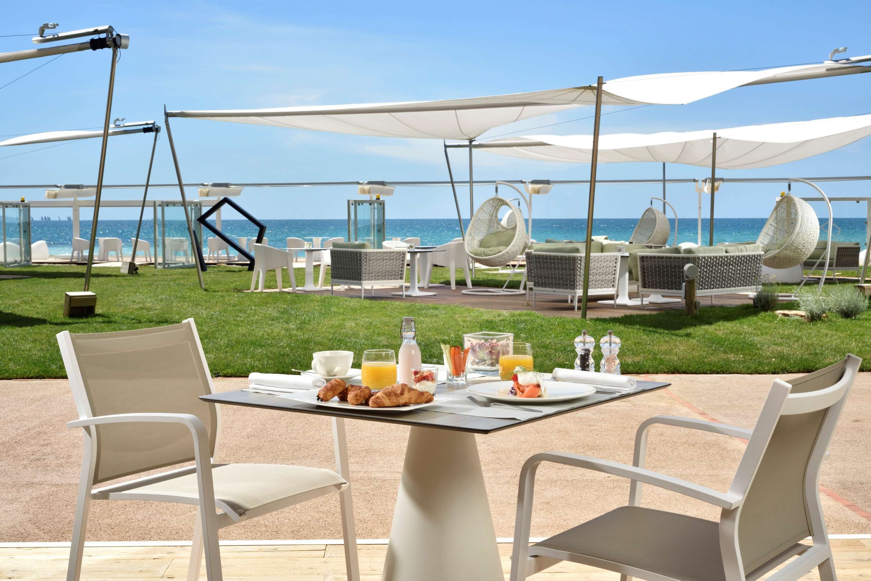 An outdoor dining area with tables and chairs, a view of the sea, and umbrellas providing shade.