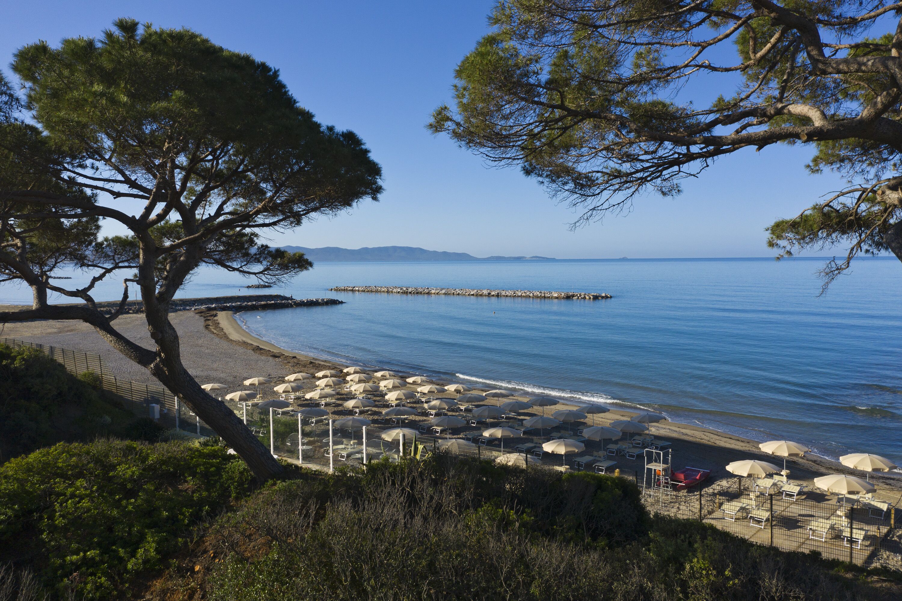 Una spiaggia con ombrelloni e lettini, un molo roccioso e un cielo azzurro intenso.