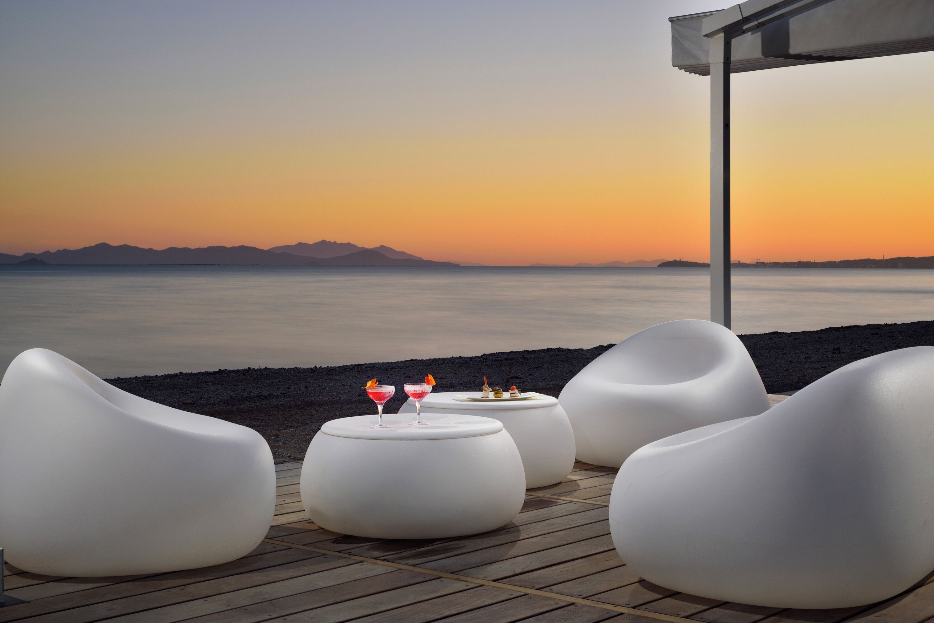 A sunset view with modern white lounge chairs and a small table with drinks on a wooden deck.