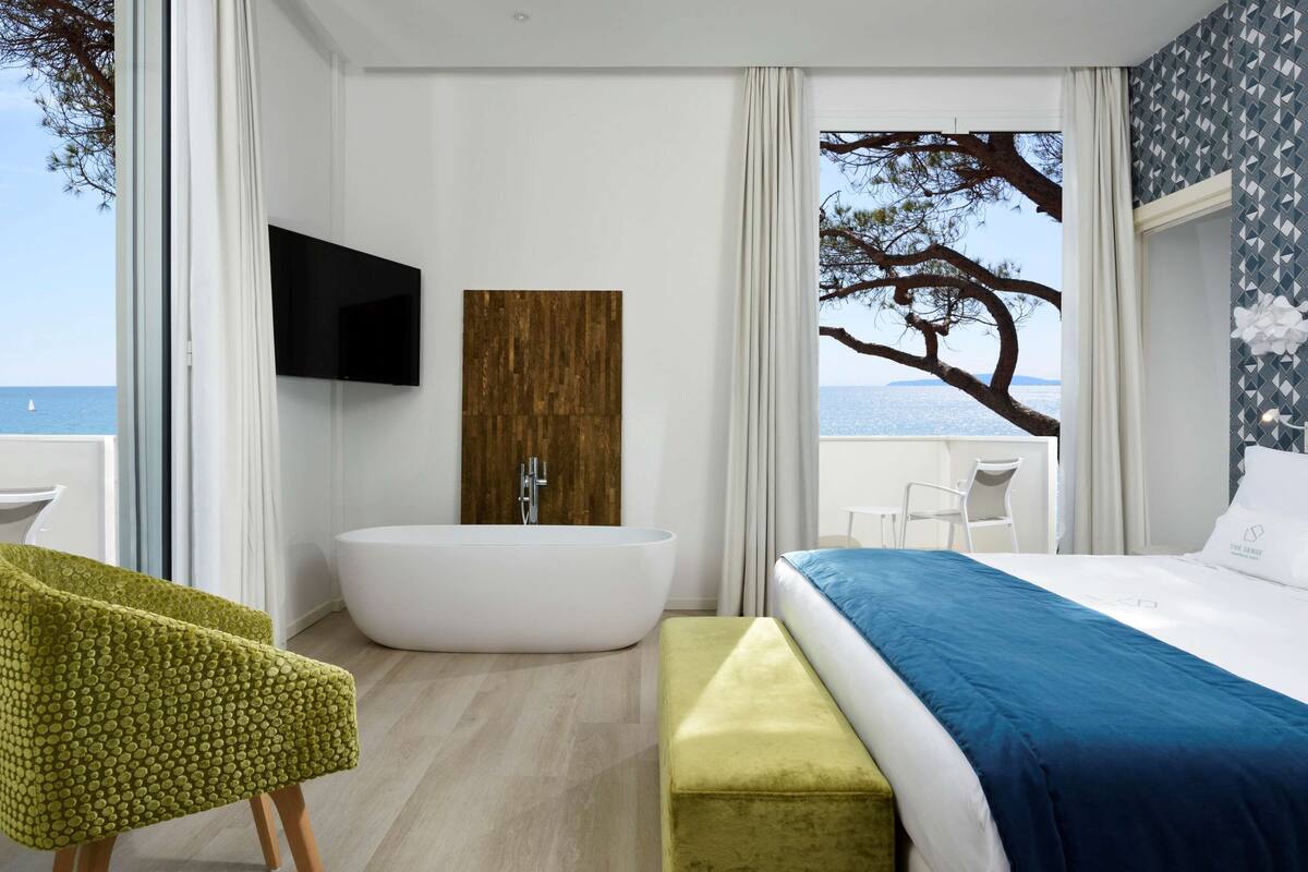 A modern bedroom with a large bed, a bathtub, a TV, and a view of the sea.