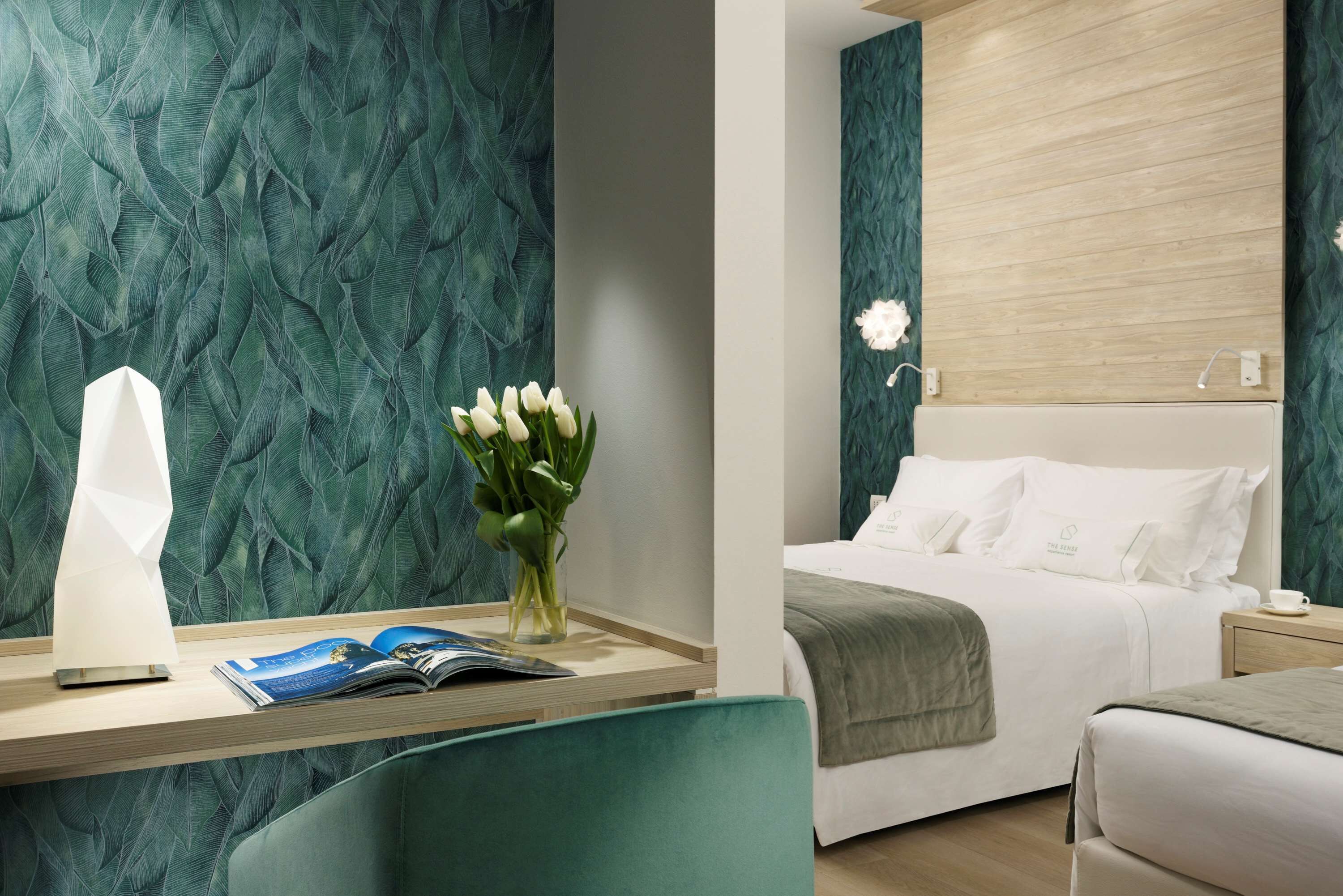 A hotel room with a teal patterned wall, a bed with white bedding, a bedside table with a lamp, a vase with white flowers, and a magazine.