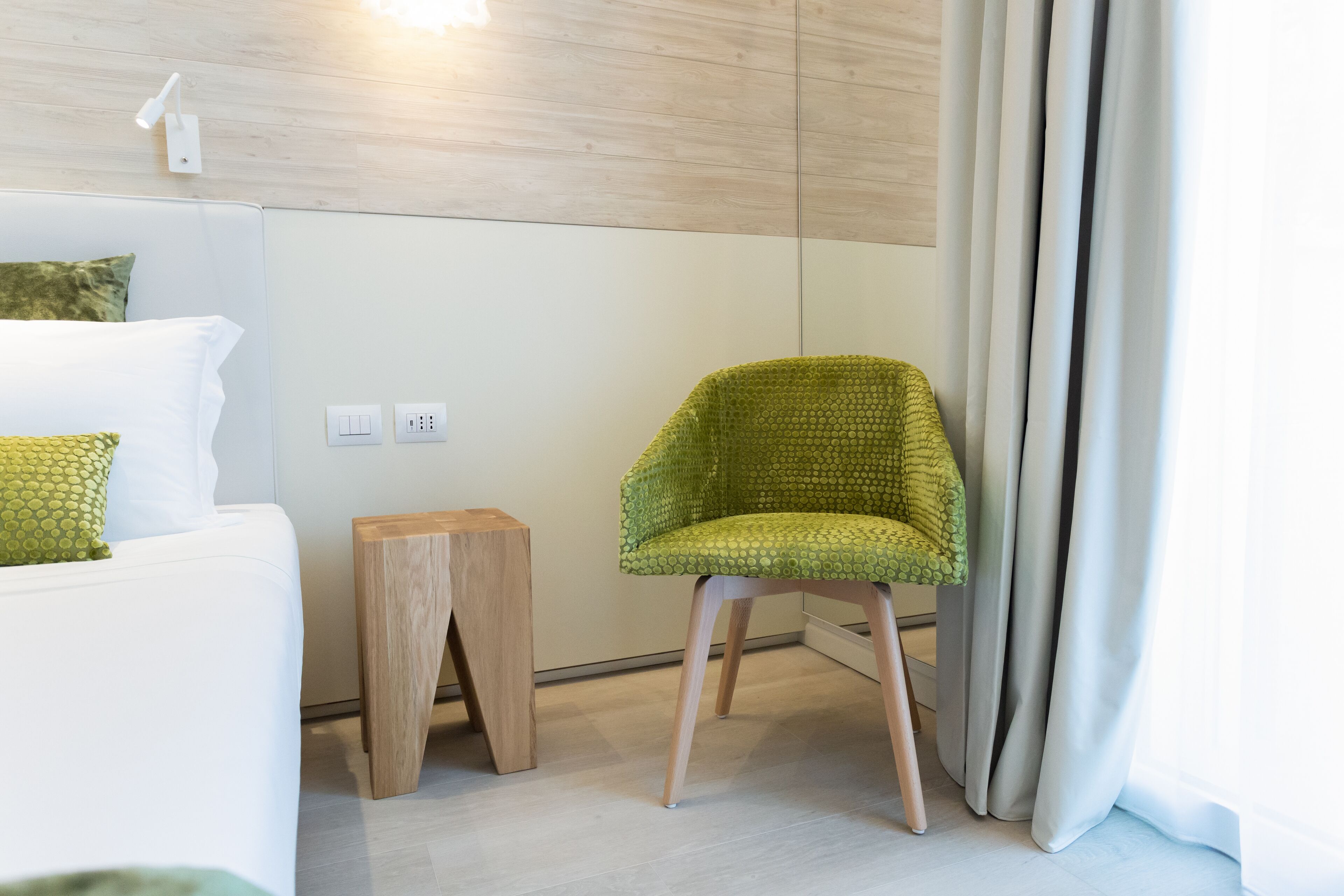 Quarto moderno com sofá branco, uma cadeira verde, um banquinho de madeira e piso em tons claros.