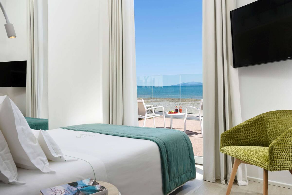 A hotel room with a bed, a green blanket, white curtains, a TV, and a view of the beach.