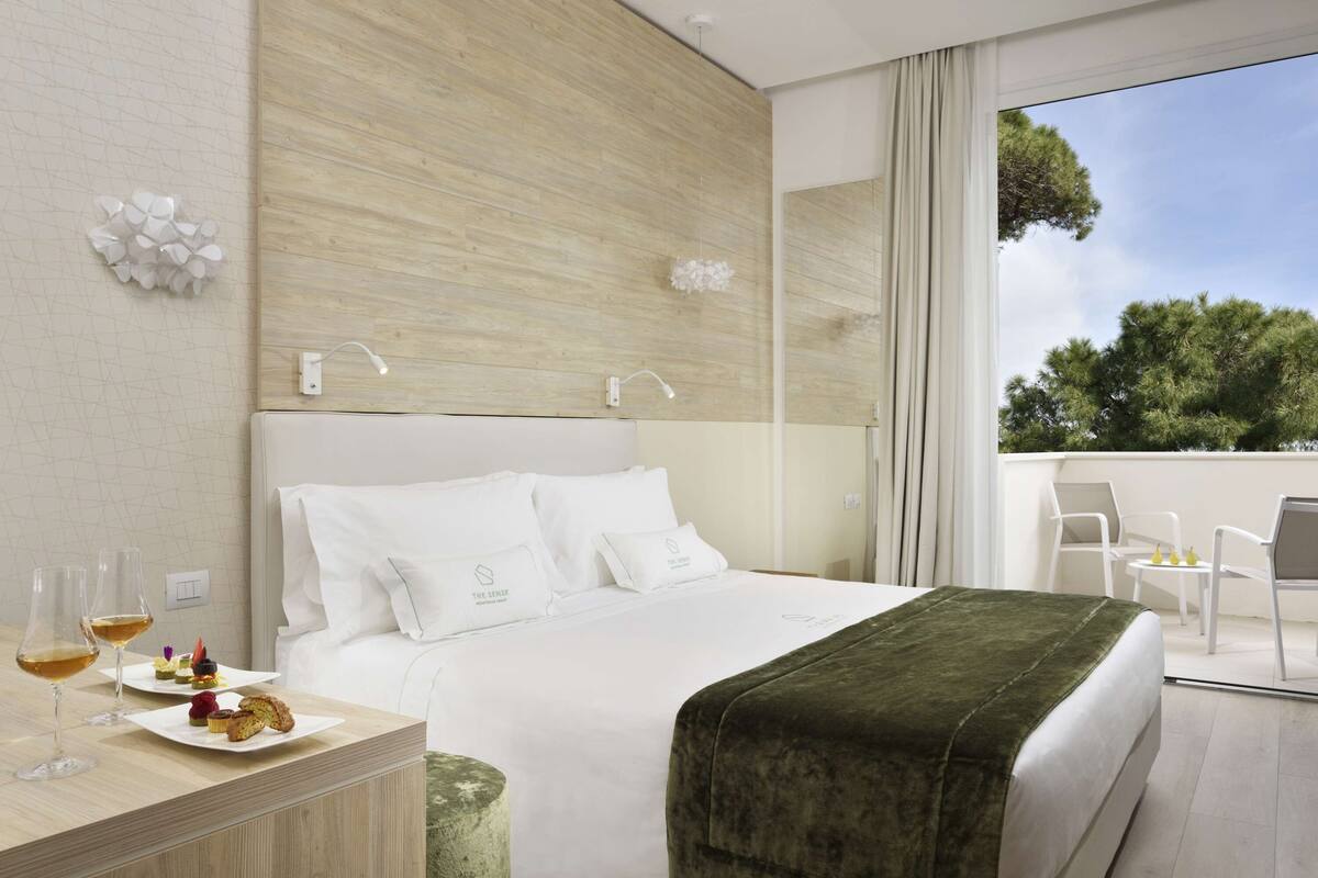 A modern hotel room with a large bed, a bedside table with glasses of wine, and a balcony with a view of trees.