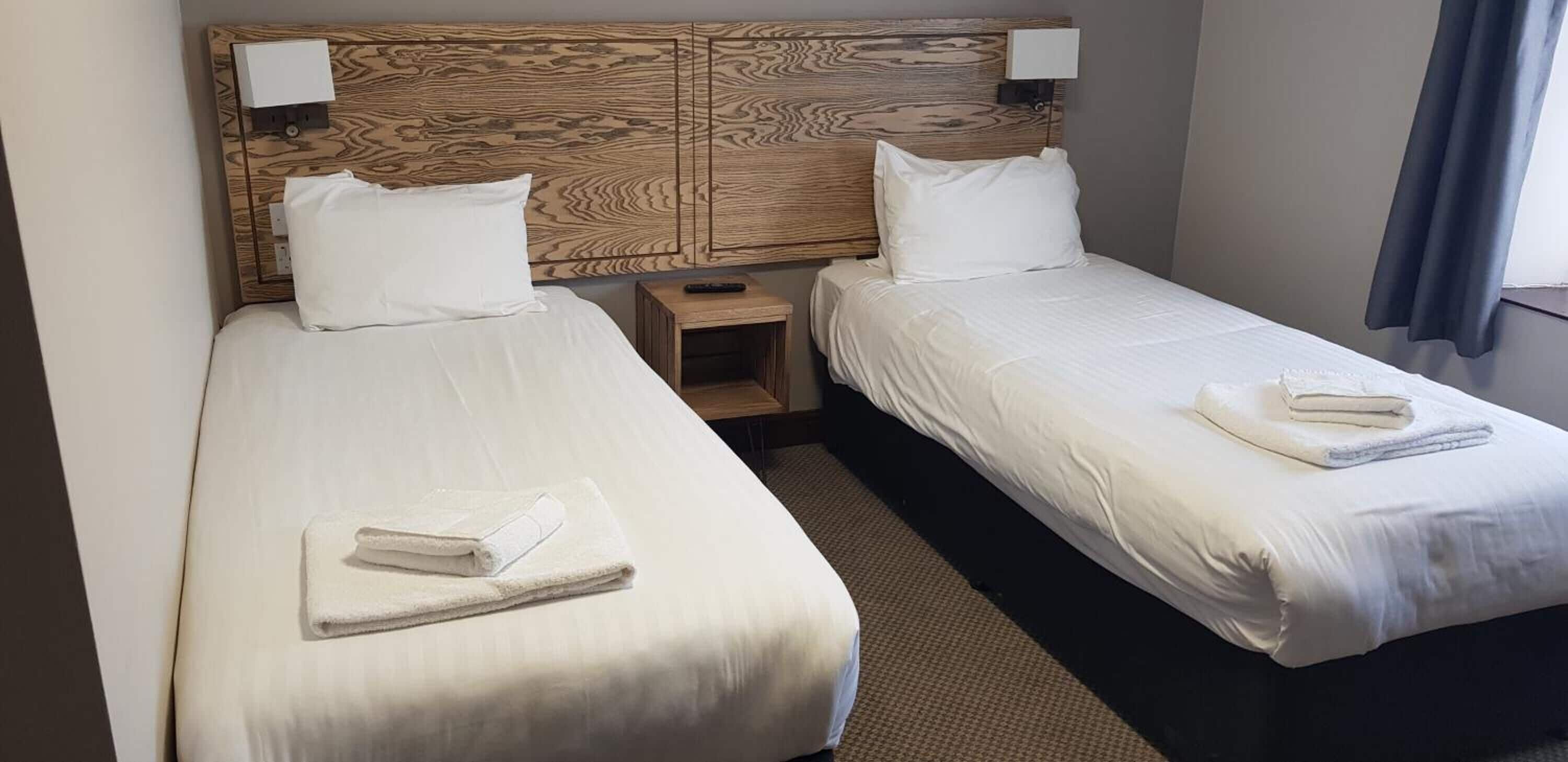 Quarto de hotel com duas camas, cada uma com lençóis e toalhas brancos, cabeceira de madeira, luminárias de cabeceira e uma pequena mesa de cabeceira de madeira.