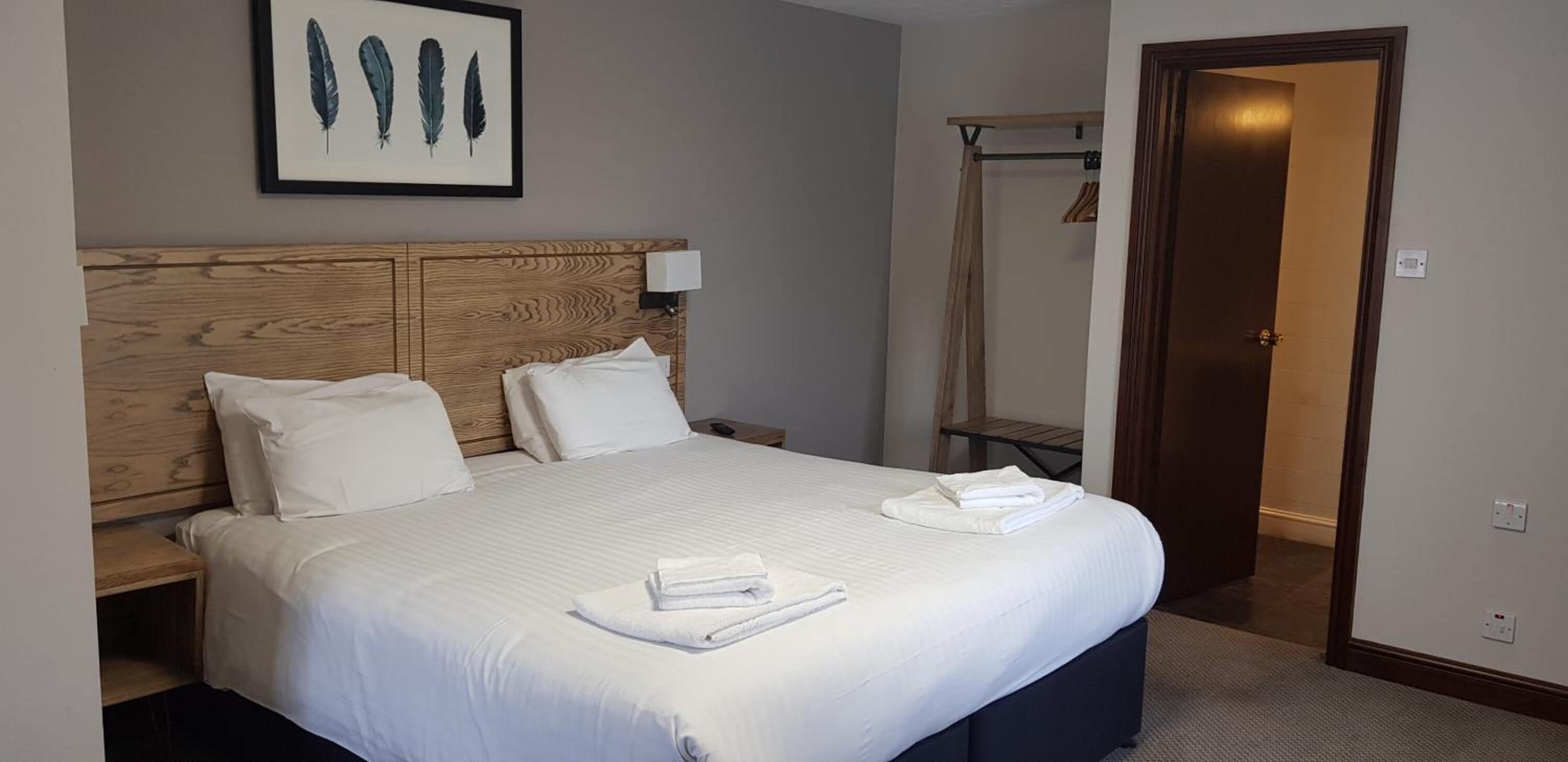 A hotel room with a large bed, wooden headboard, white bedding, a framed feather artwork, a bedside table with a lamp, and a door leading to a bathroom.