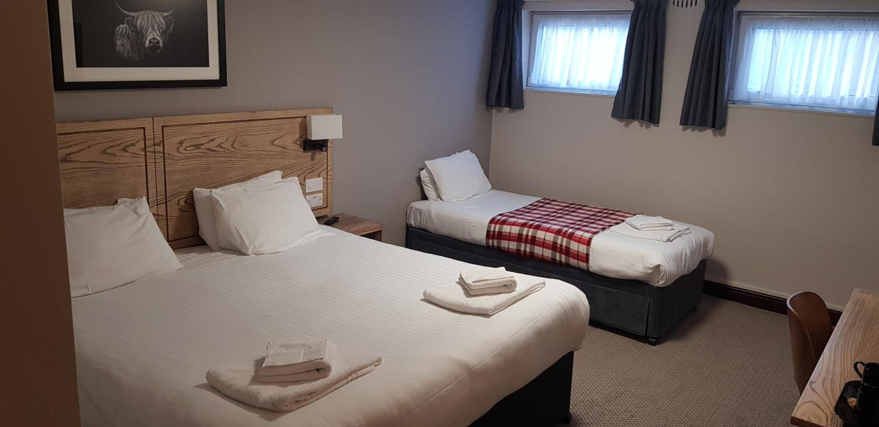 A hotel room with two beds, a wooden headboard, a small table, and a window with curtains.