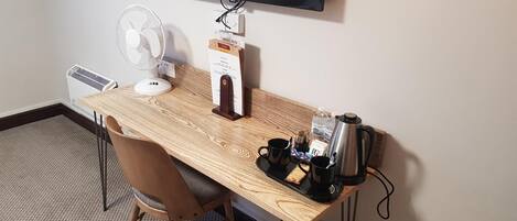 Escritorio de madera con un ventilador, una cafetera y una bandeja con dos tazas y una galleta.