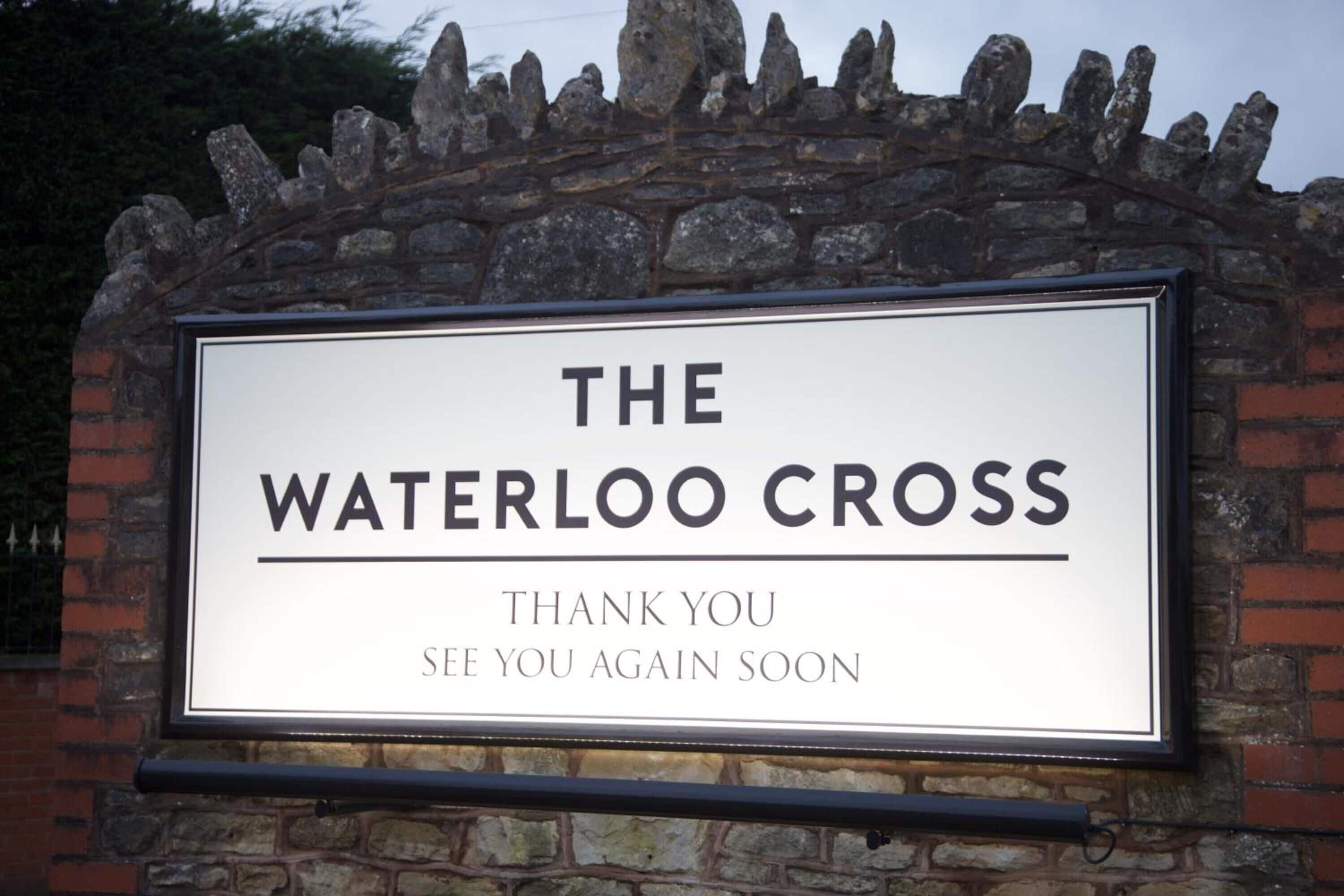 Un letrero con el texto 'THE WATERLOO CROSS' y el mensaje 'THANK YOU SEE YOU AGAIN SOON' en una pared de piedra.