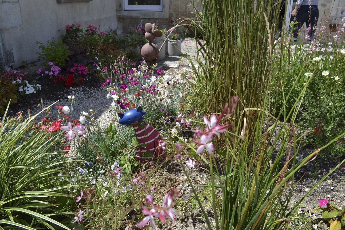 Un jardín con diversas plantas y flores, una estatua y una persona caminando al fondo.
