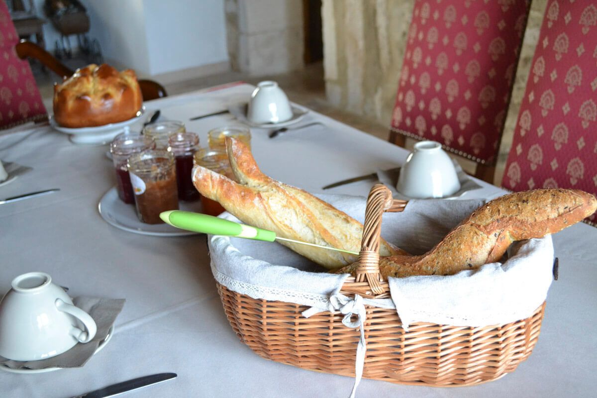 Uma mesa de jantar com uma cesta de baguetes, um pão e vários pequenos potes de geleia.