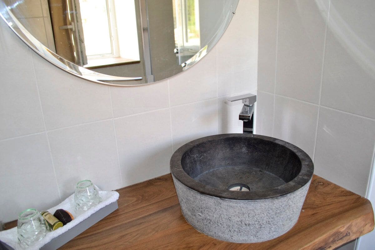 A bathroom with a stone basin sink, a wooden countertop, a mirror, and a shelf with toiletries.