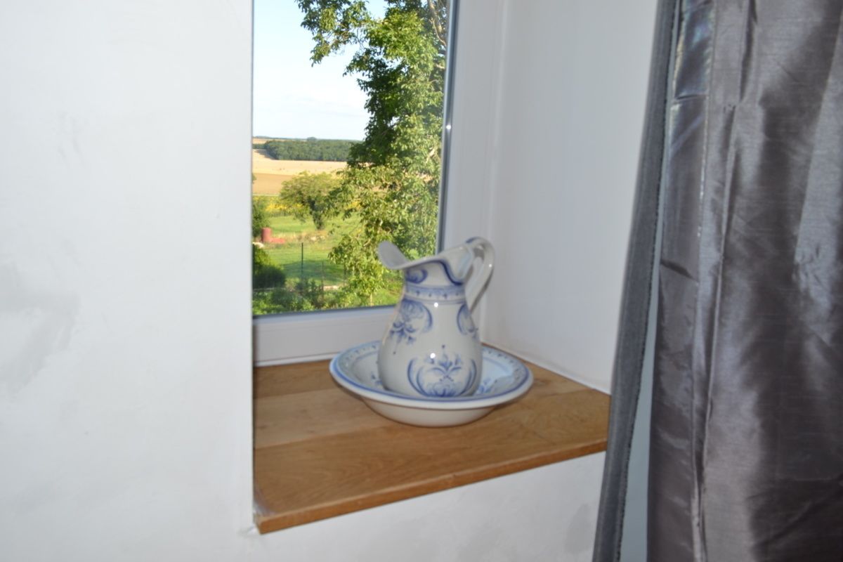 A ceramic pitcher on a wooden shelf by a window with a view of greenery and fields.