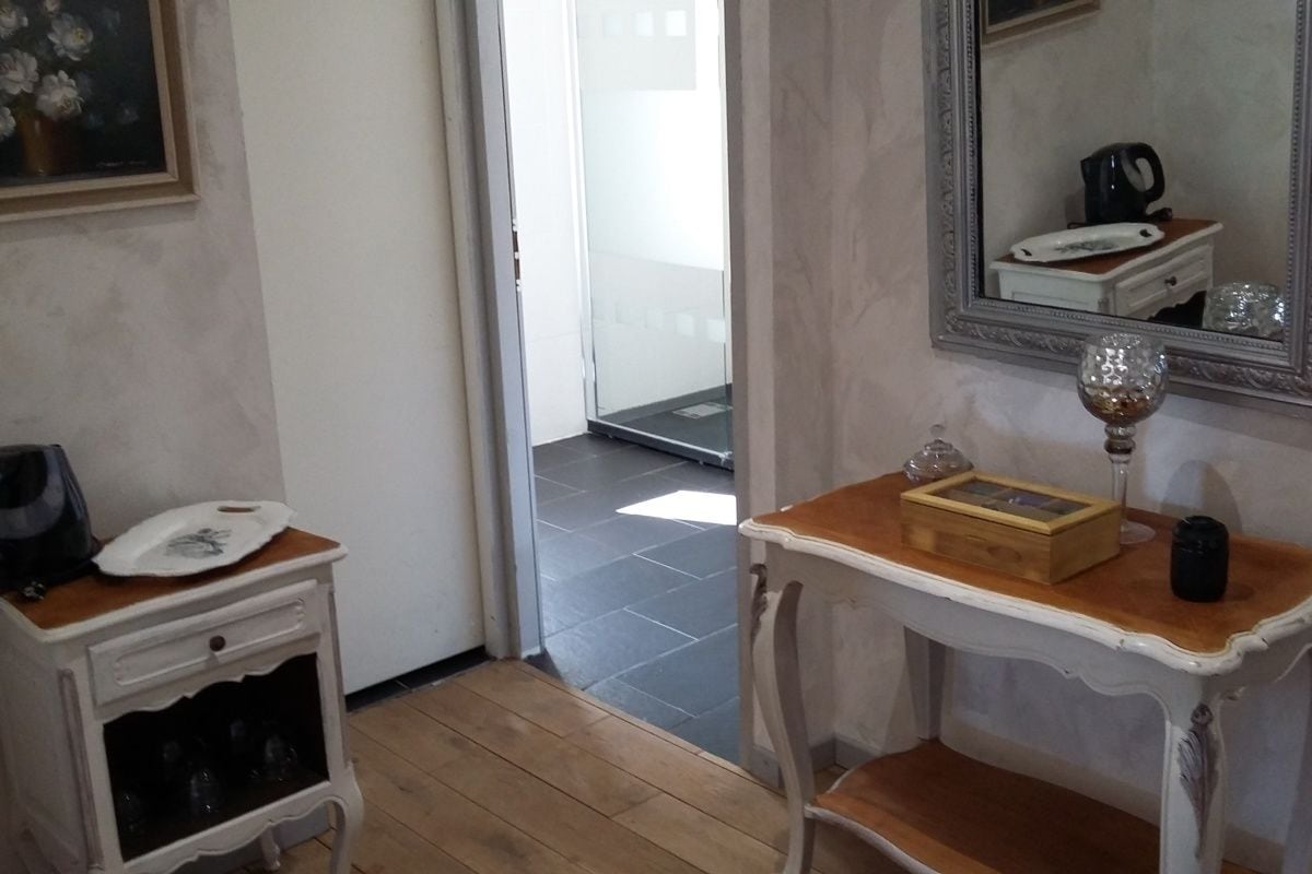 Una stanza con pavimento in legno, una cassettiera bianca con telefono e un bicchiere da vino, un tavolino con una scatola e un bicchiere, e uno specchio appeso al muro.