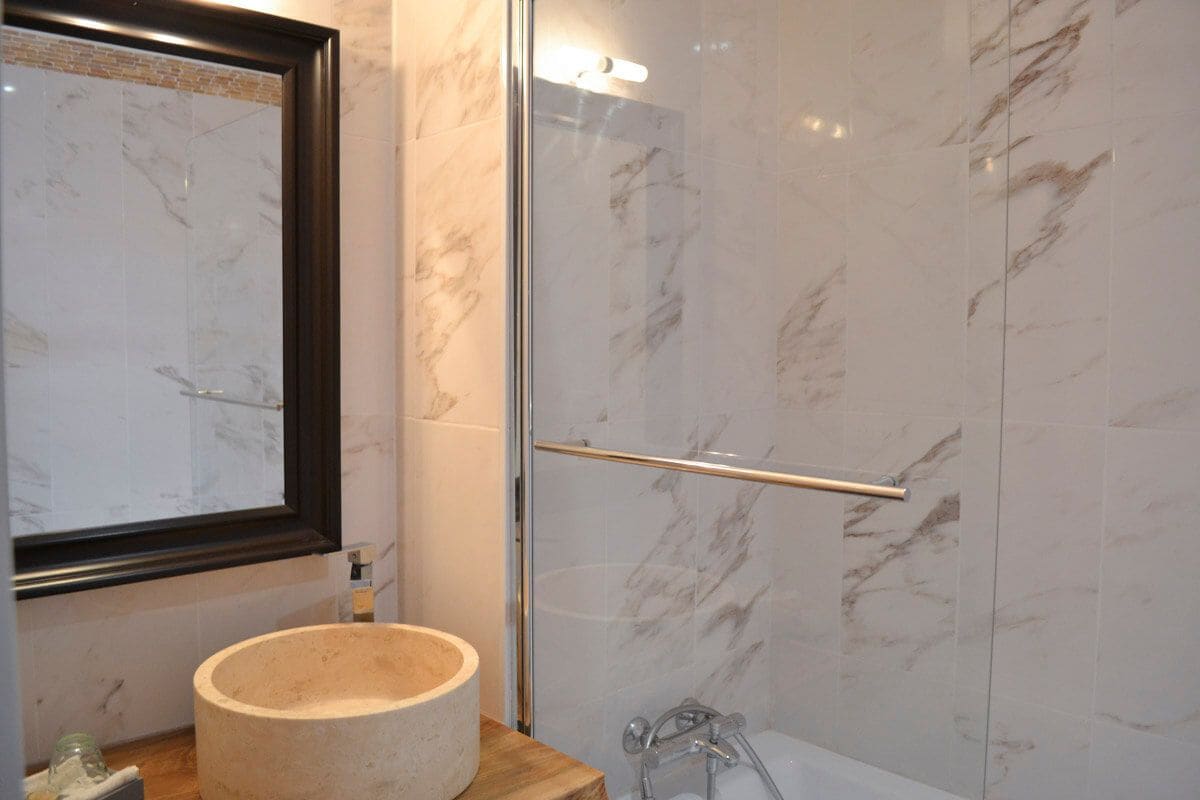 Un baño con pared de azulejos de mármol, ducha con cerramiento de vidrio, tocador de madera con lavamanos redondo y espejo enmarco.