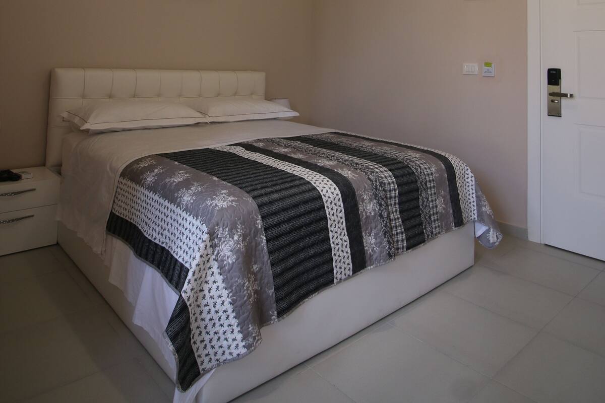A bedroom with a bed, a bedside table, a nightstand, a wall-mounted light, and a door.