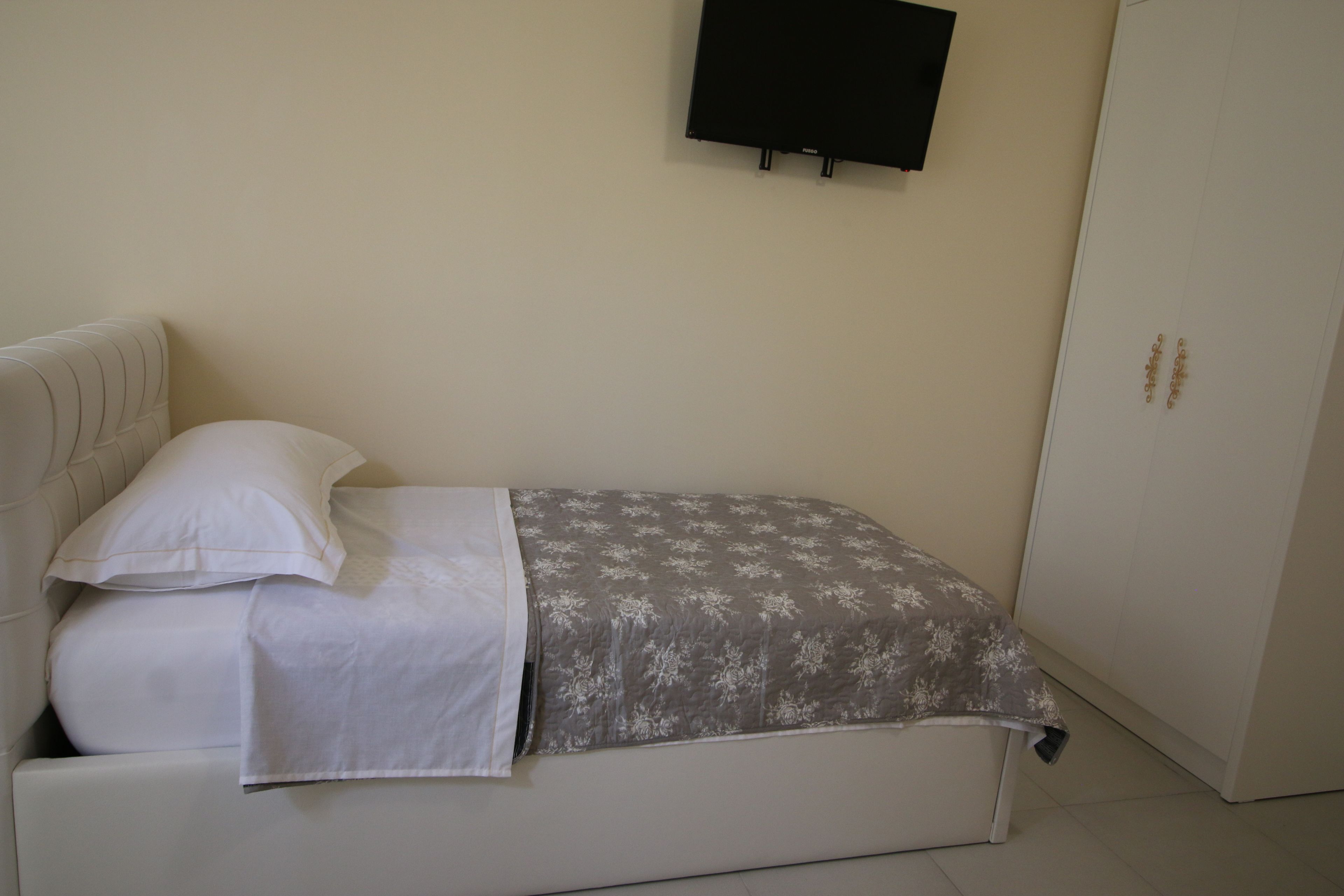 Ein Einzelbett mit weißem Kopfteil und Kissen, eine graue floral gemusterte Bettdecke und ein schwarzer Fernseher an der Wand darüber.